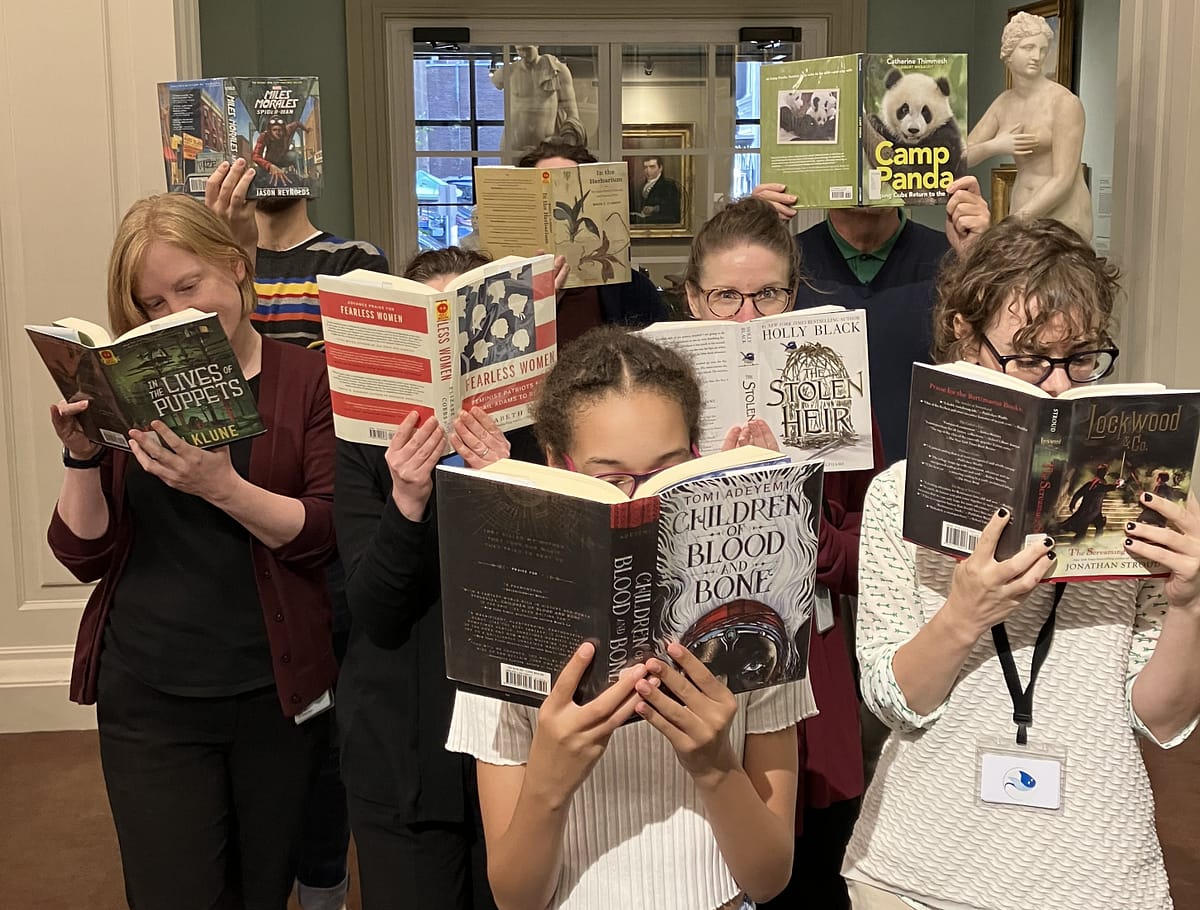 Six staff members holding books up in front of their faces.